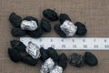 A collection of small owl pellets, mostly black in color, with a few wrapped in aluminum foil, displayed on a beige background with a measuring tape for scale.