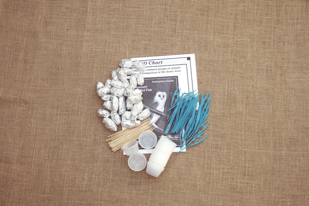 Educational kit for owl pellet dissection, including wrapped owl pellets, an instructor's guide, a bone ID chart, wooden dissecting probes, and plastic forceps, displayed on a burlap-like surface.